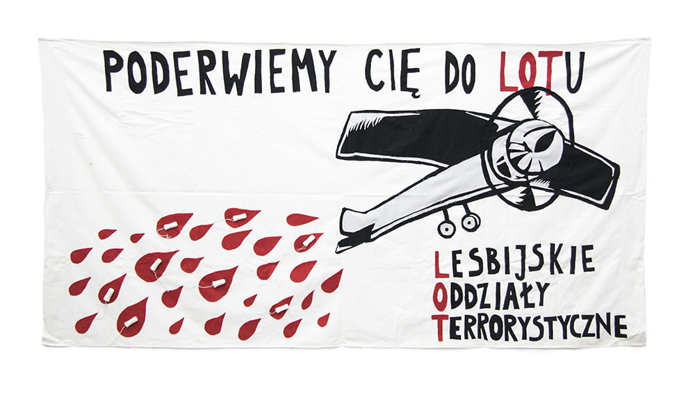 Zdjęcie. Biały transparent przedstawiający samolot i krople krwi z tamponami. Napis "Poderwiemy cię do lotu. Lesbijskie Oddziały Terrorystyczne"