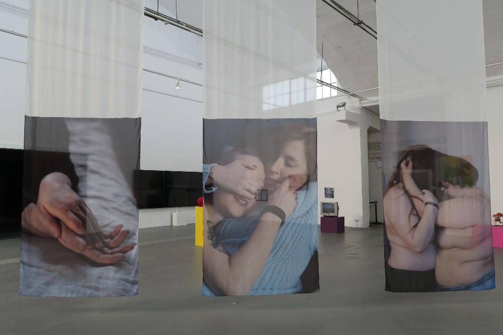 Zdjęcie. W sali wiszą trzy tkaniny z wydrukiem zdjęć przytulających się kobiet.
