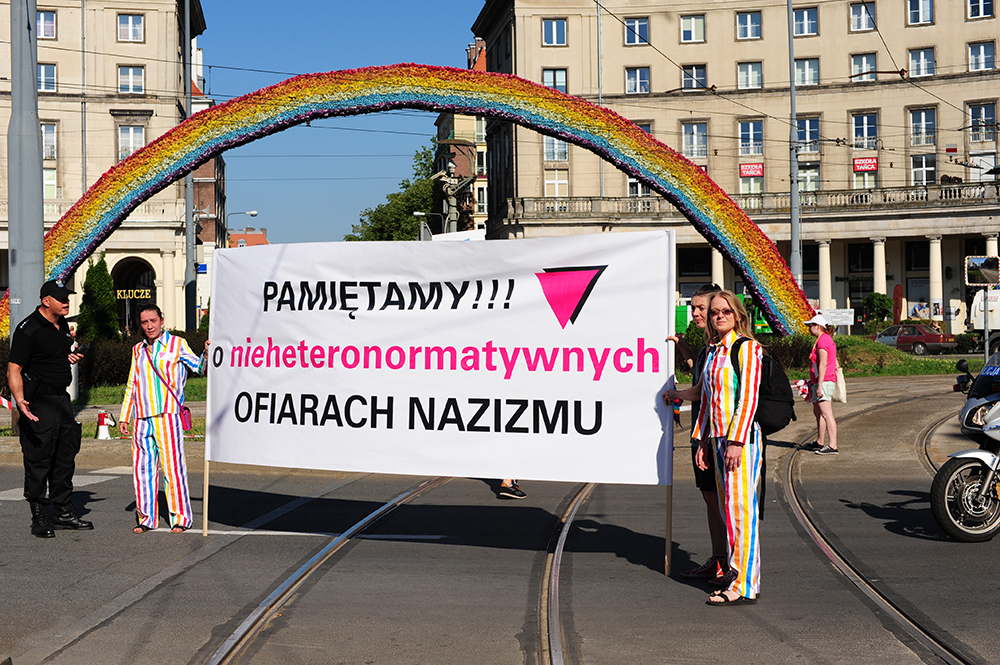 Zdjęcie. Osoby w kolorowych pasiakach trzymają biały transparent z napisem "Pamiętamy o nieheteronormatywnych ofiarach nazizmu". W tle tęcza.