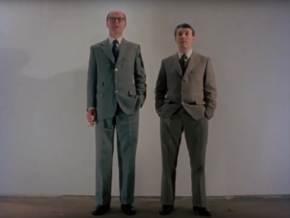 Kadr z filmu. Dwóch mężczyzn w garniturach stoi obok siebie. W tle biała ściana.