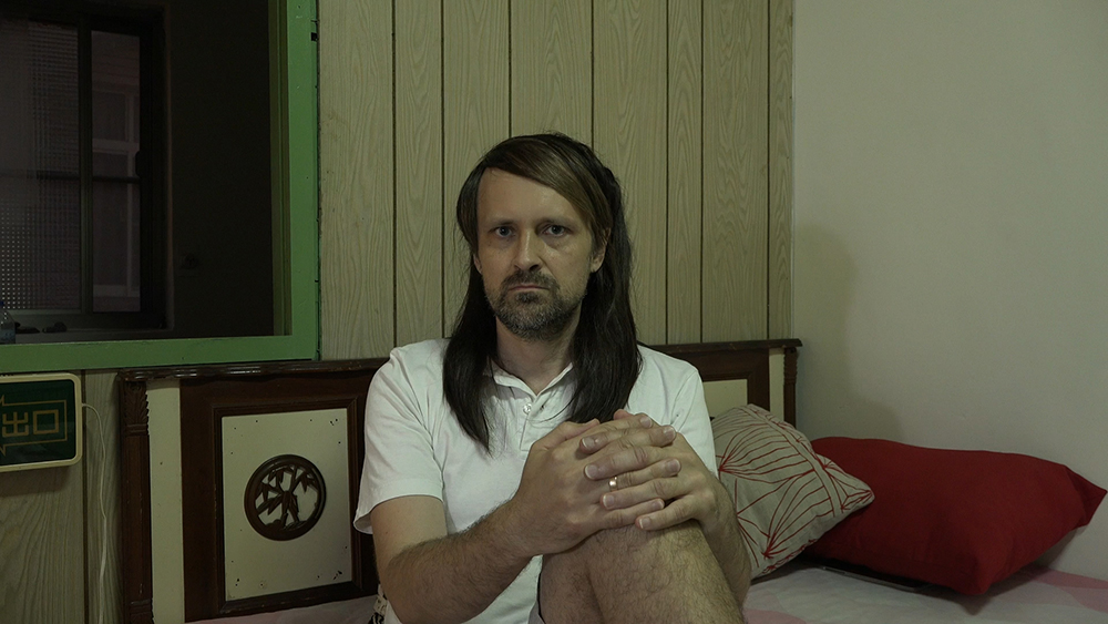 Zdjęcie. Mężczyzna z długimi włosami i brodą, w białej koszulce siedzi na łóżku. 
