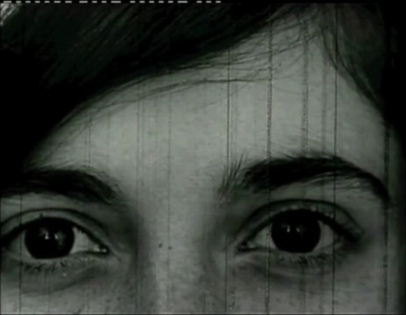 Kadr z czarno-białego filmu. Zbliżenie na twarz osoby z ciemnymi oczami i włosami.