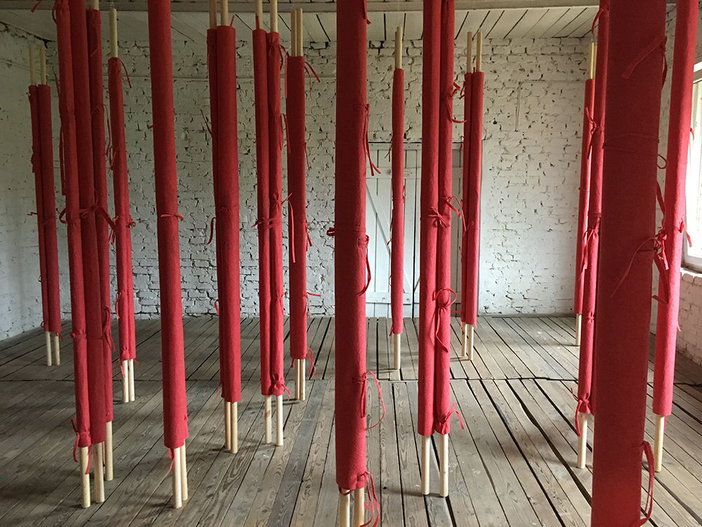 Zdjęcie. Wnętrze o białych, ceglanych ścianach w którym stoją wysokie, drewniane paliki obwiązane czerwonym sznurkiem.